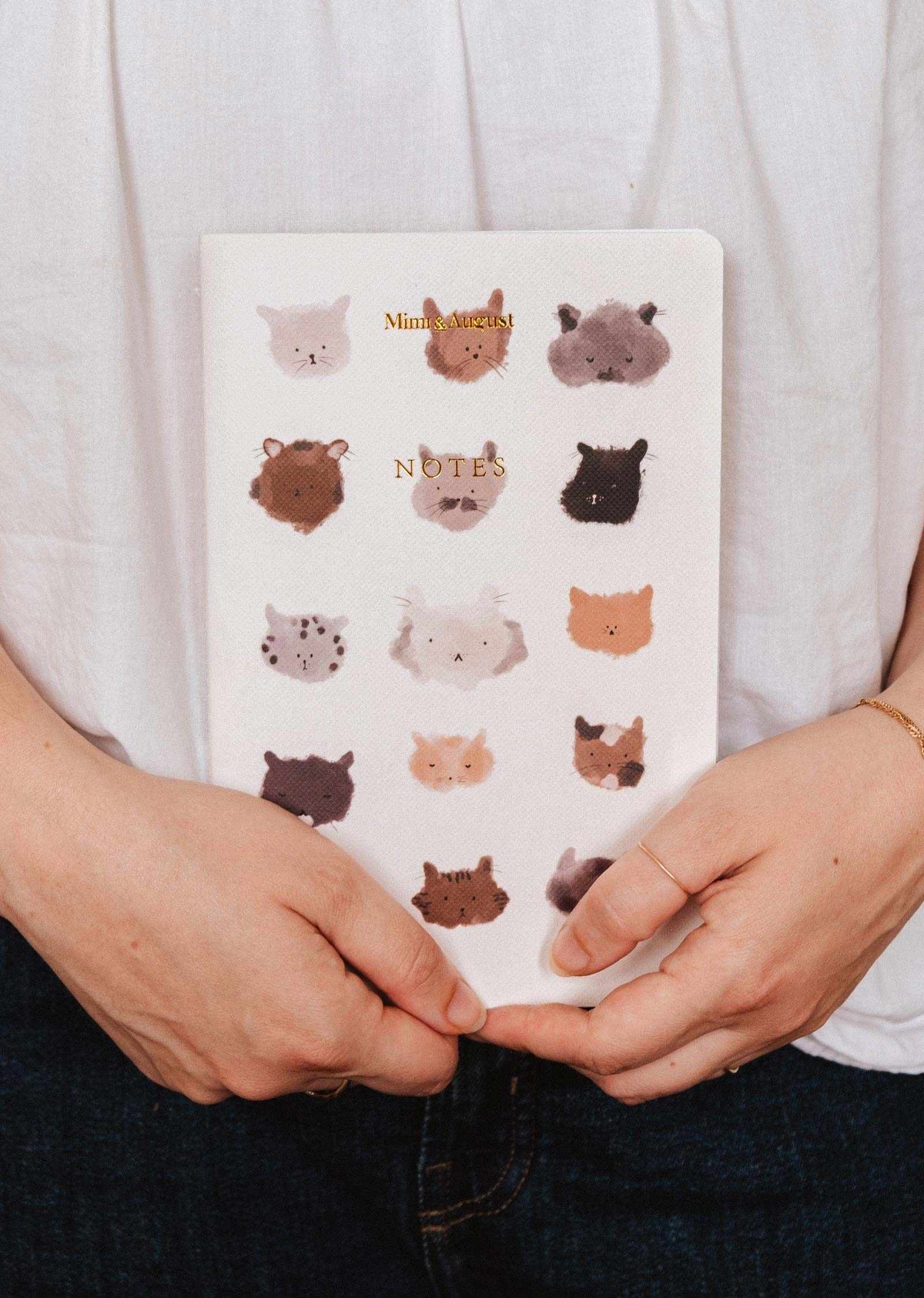 Someone holds The Purrfect - Notebook by Mimi & August, a white notebook with illustrated animal faces in a grid on the cover—ideal for cute stationery lovers.