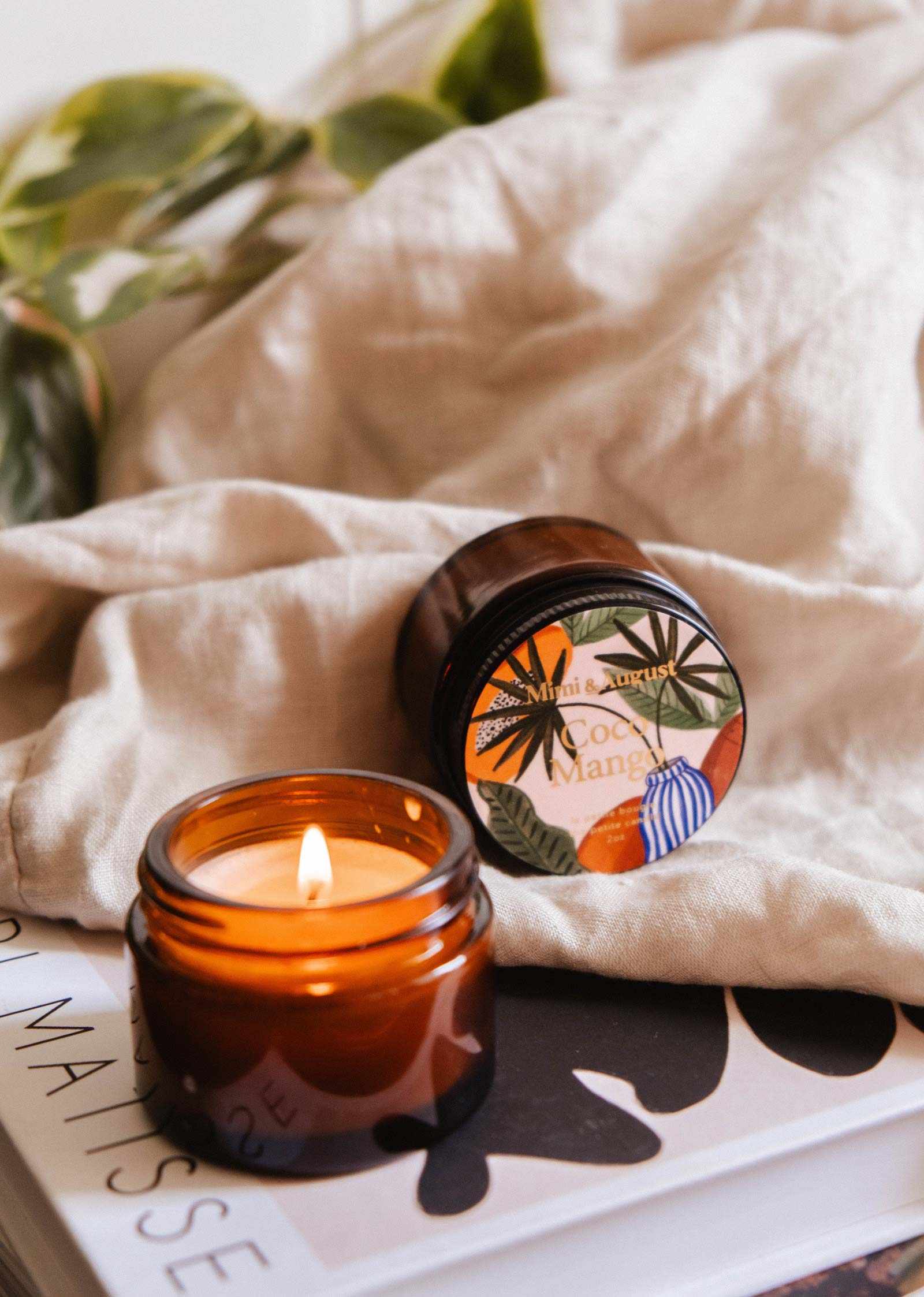 A lit Mimi & August Coco Mango - 2 oz Soy Candle in an amber glass jar fills the air with a tropical scent. The reusable candle sits on a book by its lid, with a beige cloth and green leaves in the background.