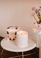 The Bloom – Reusable Soy Candle by Mimi & August, in a ribbed holder, glows on a round marble table beside a floral box and pink flowers in a vase.