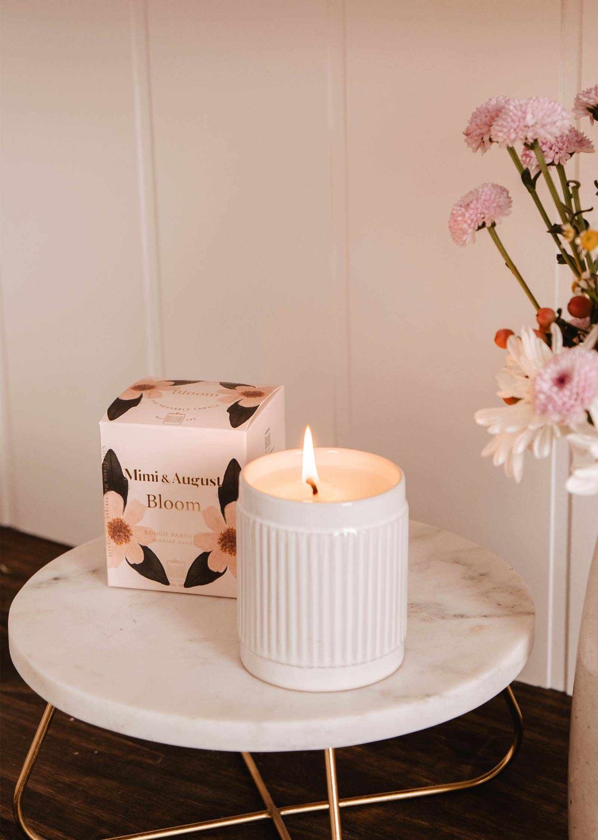 La bougie de soya réutilisable Bloom de Mimi & August, dans un bougeoir cannelé, brille sur une table ronde en marbre à côté de sa boîte florale et de fleurs roses dans un vase.