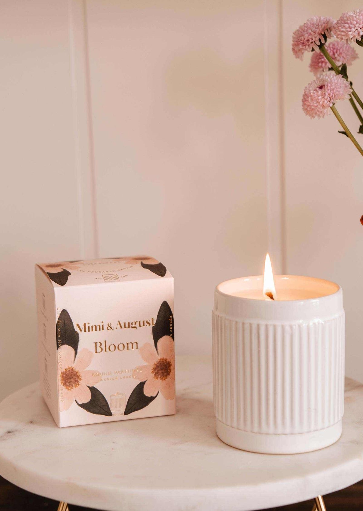 Une bougie de soya réutilisable Bloom de Mimi & August allumée au parfum d'orange et canneberge repose sur une surface en marbre à côté d'une boîte florale « Bloom », avec des fleurs roses dans un vase en arrière-plan.