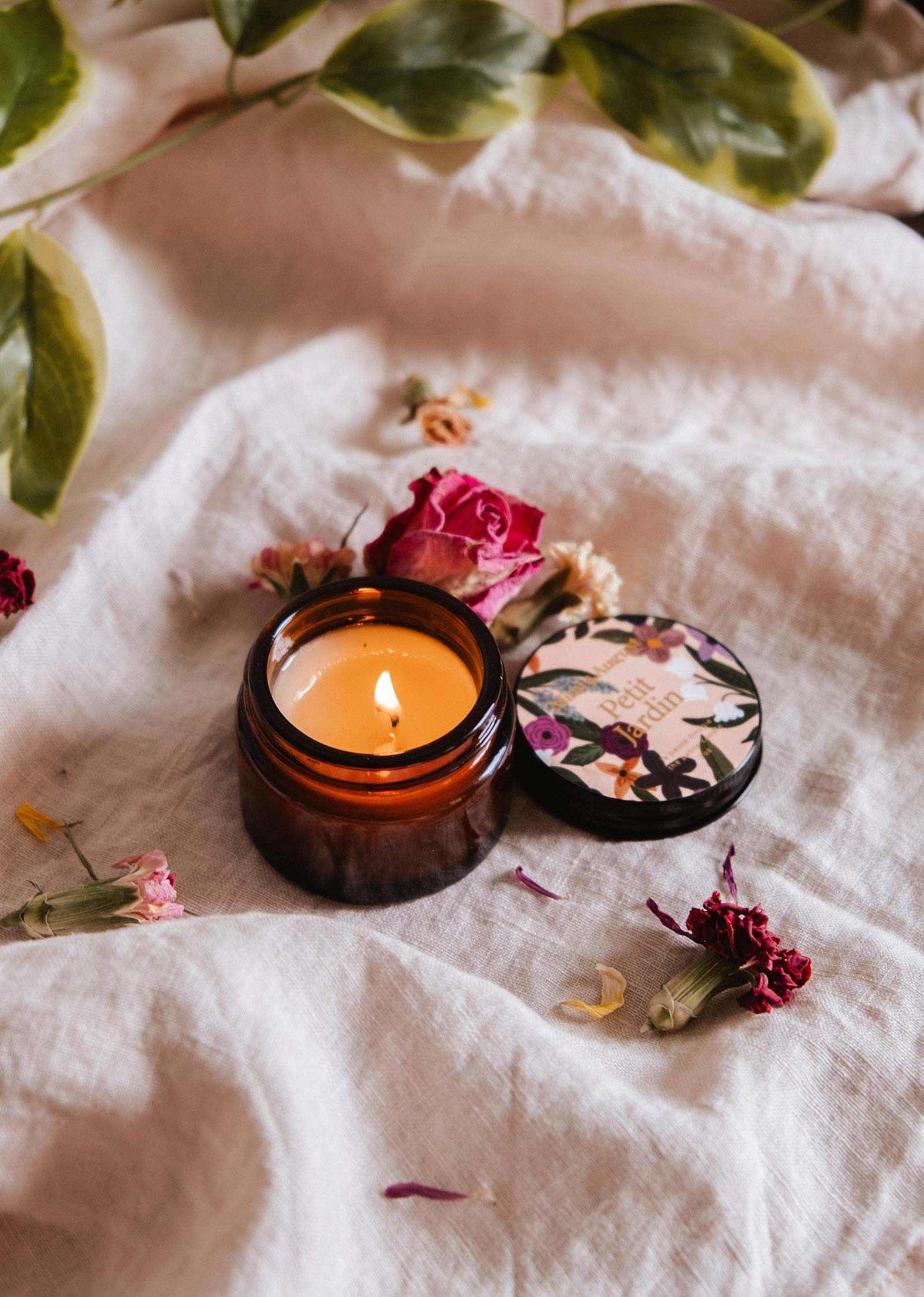 La bougie de soya Petit Jardin 2 oz de Mimi & August est une bougie réutilisable coulée à la main dans un petit pot floral. Ouverte et allumée, elle repose sur un tissu blanc avec des fleurs séchées et des feuilles vertes, ajoutant du charme à toute pièce. Faite à Montréal.
