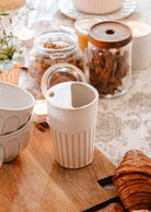The Ribbed To-Go Cup from Mimi & August is stacked alongside a ceramic pitcher on a wooden board with a croissant, glass jars of granola and dried fruit, and small lit candles atop a patterned tablecloth.