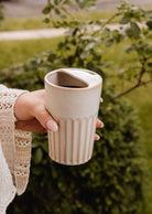 A hand holding The Ribbed To-Go Cup by Mimi & August with a partially open lid, outdoors near green bushes.