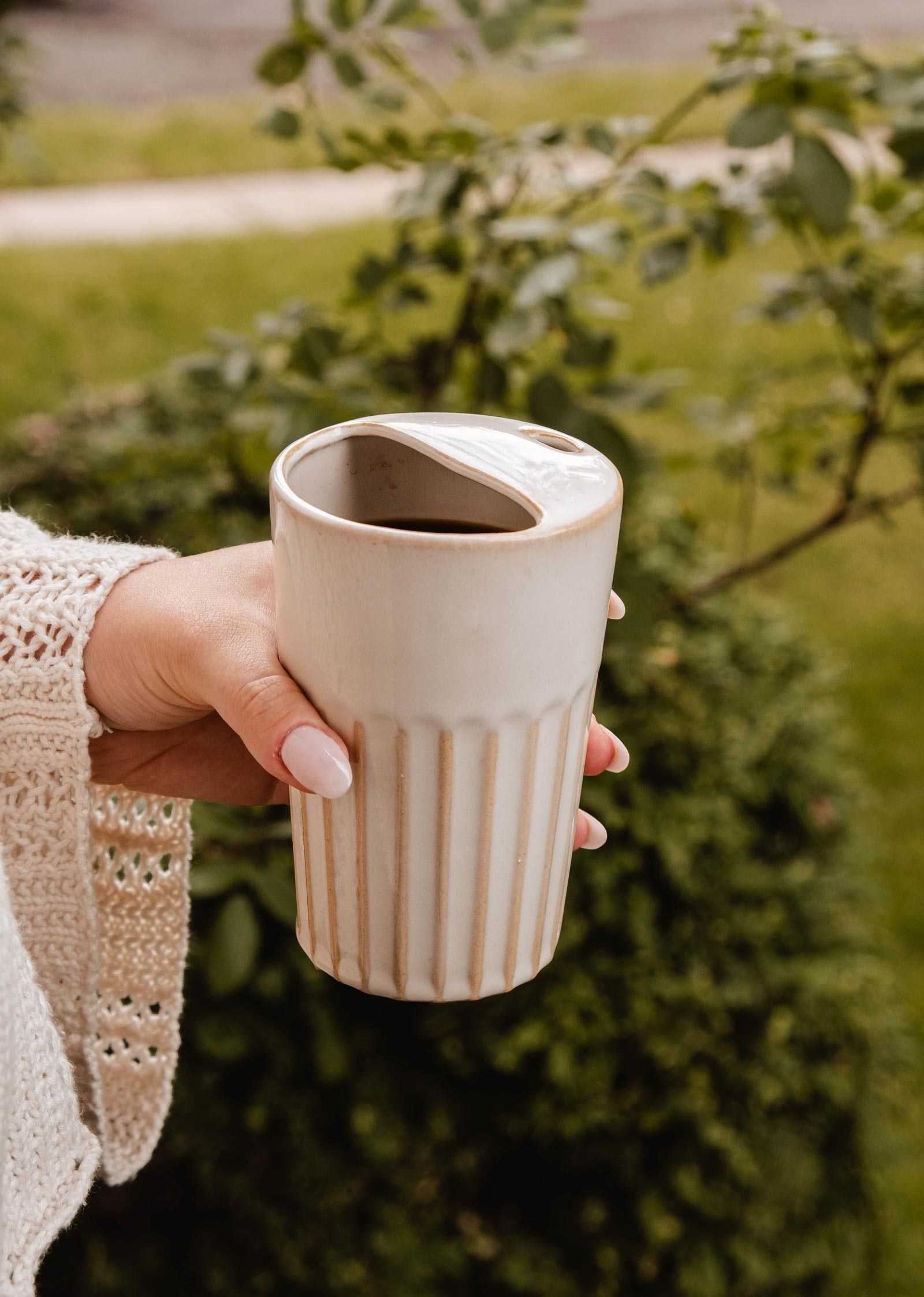 A hand holding The Ribbed To-Go Cup by Mimi & August with a partially open lid, outdoors near green bushes.