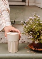 A person in a cream sweater holds the Ribbed To-Go Cup by Mimi & August next to a potted flowering plant on painted wooden steps.