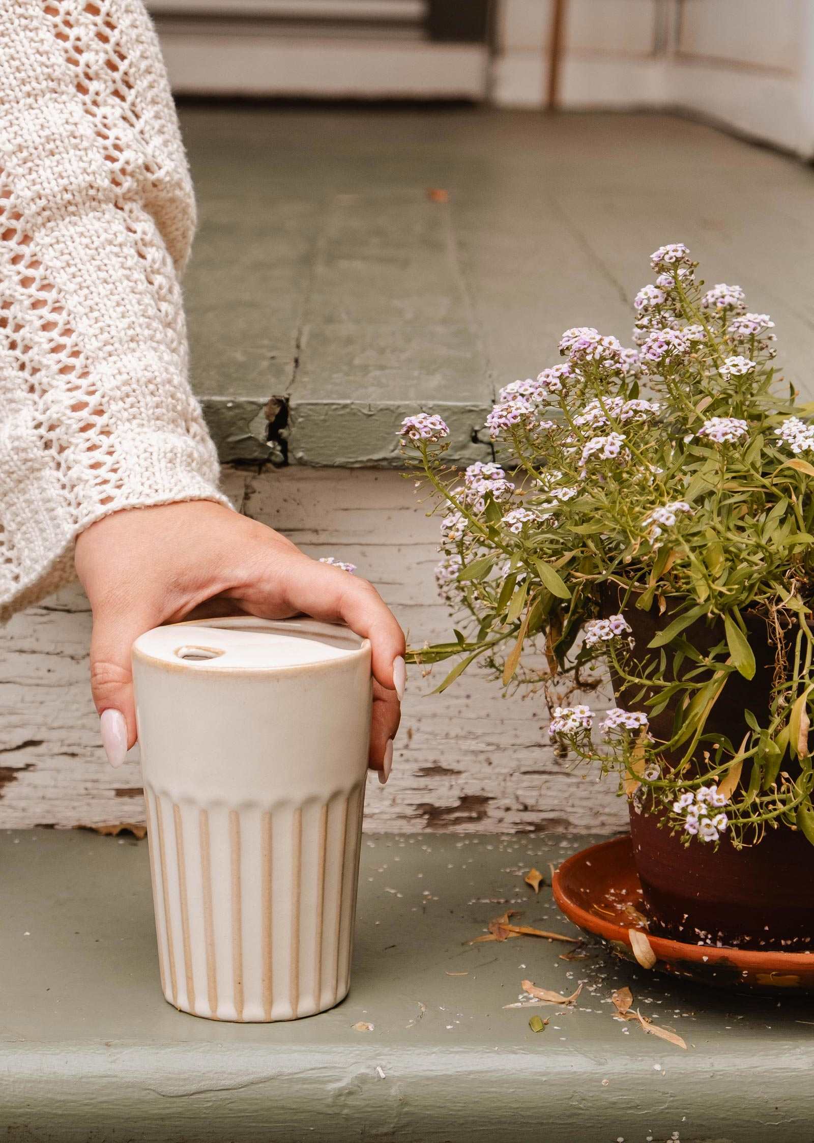A person in a cream sweater holds the Ribbed To-Go Cup by Mimi & August next to a potted flowering plant on painted wooden steps.