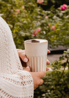 A person wearing a knitted sweater holds the Mimi & August Ribbed To-Go Cup outside, surrounded by green plants and pink flowers.