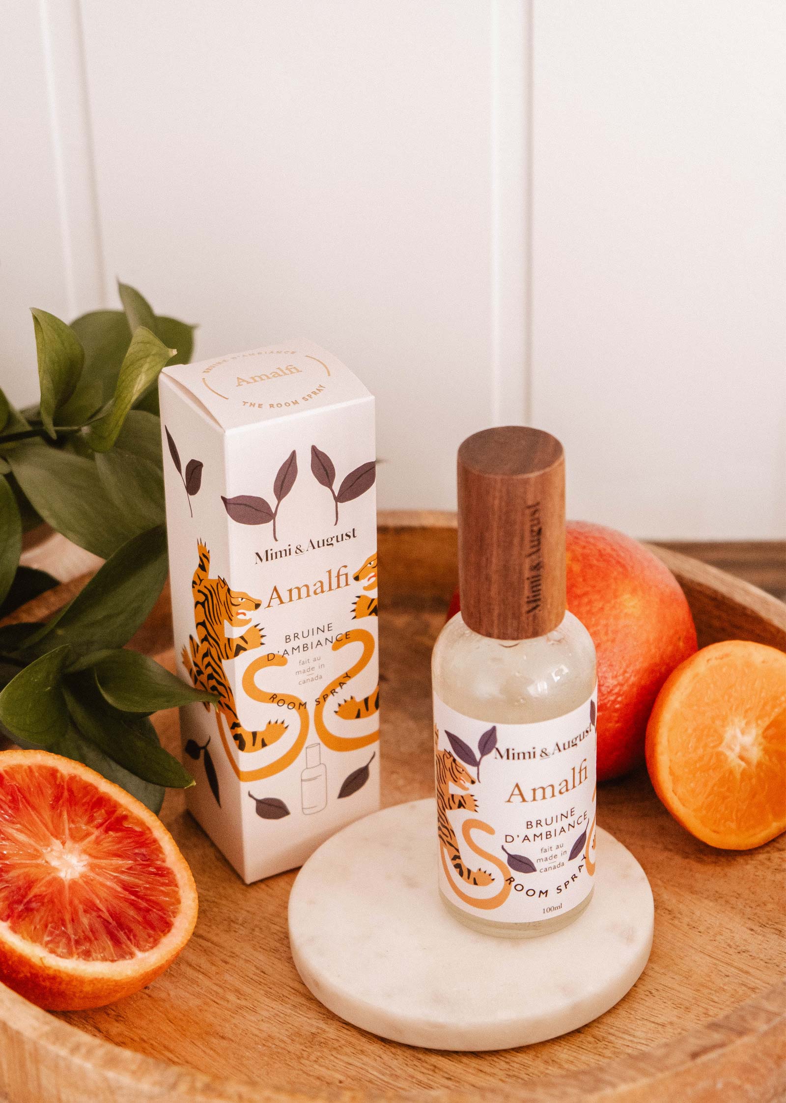 A bottle and box of Mimi & August Room Spray - Amalfi sit on a wooden tray with blood oranges and green leaves, evoking the citrusy sparkle of a Mediterranean escape.