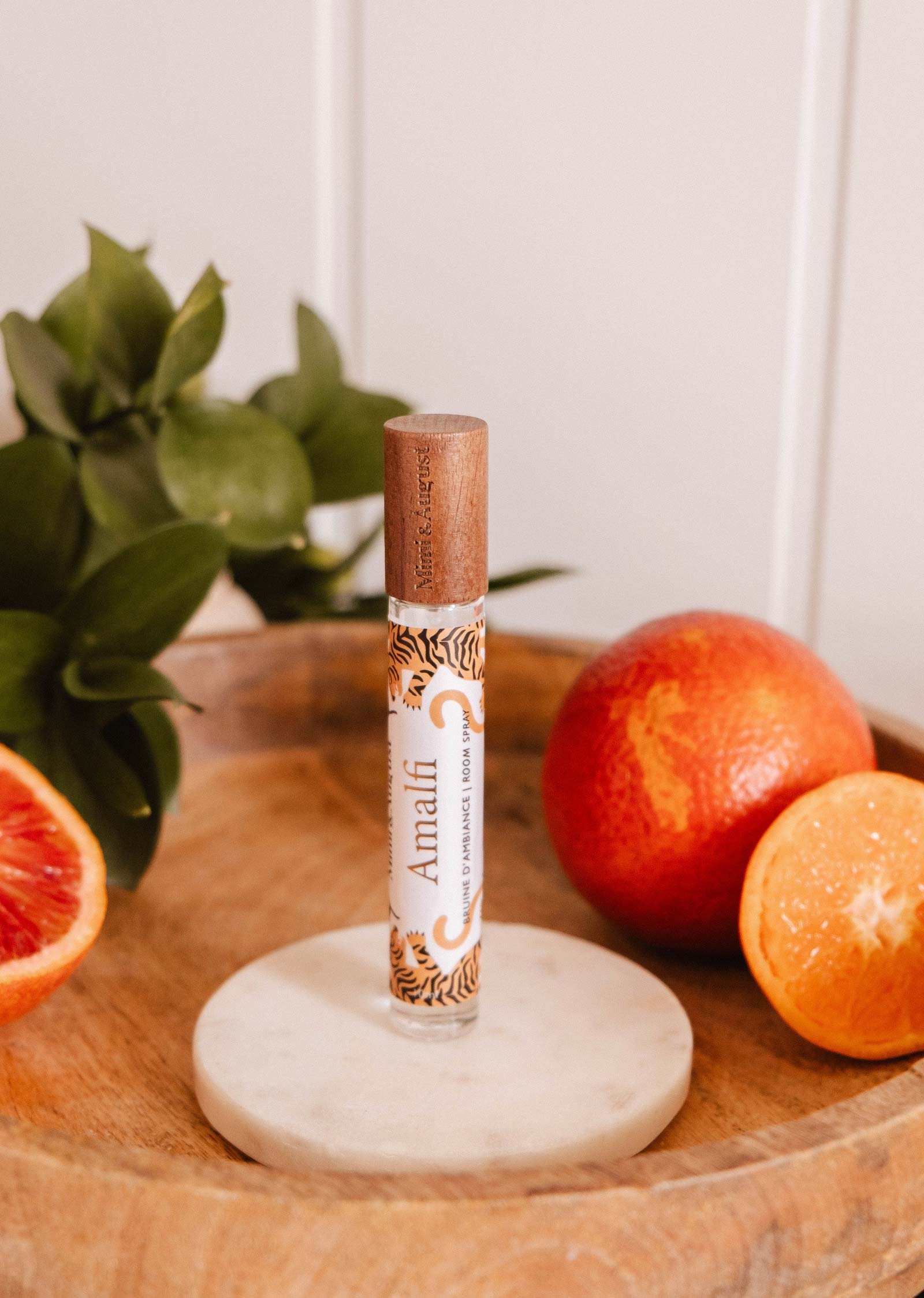 A bottle of Mimi & August Room Spray - Amalfi stands upright on a round marble coaster with blood oranges and green leaves, evoking the Mediterranean fragrance and citrus sparkle of this Amalfi-inspired scent.