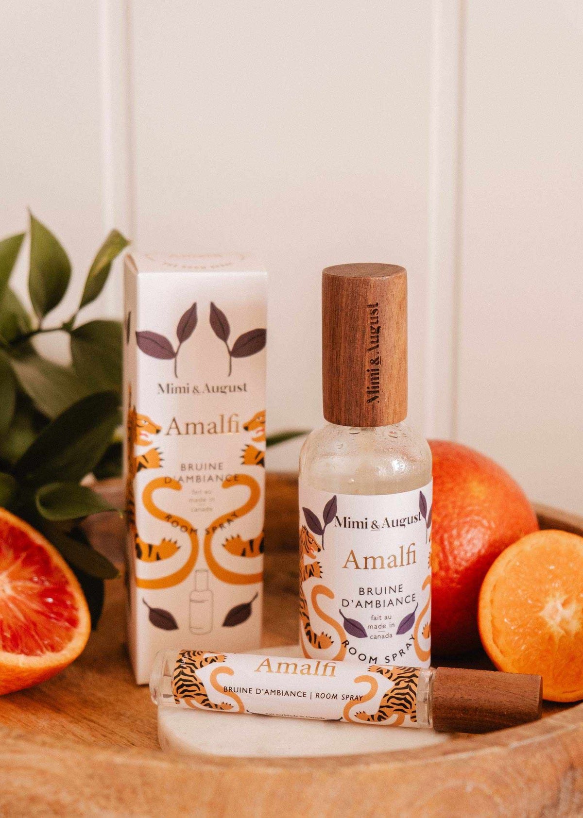 Une bouteille et une boîte de bruine d'ambiance Amalfi de Mimi & August sont présentées sur une surface en bois avec des oranges coupées et des feuilles vertes, évoquant un parfum d'agrumes rafraîchissant pour un espace d'inspiration méditerranéenne.