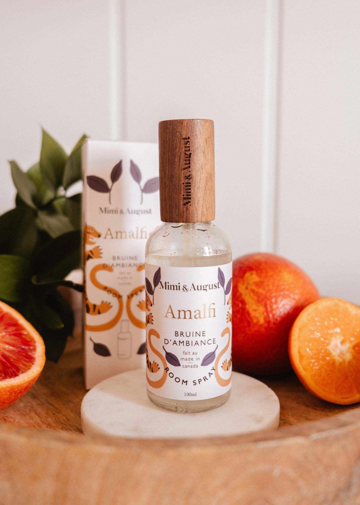 Une bouteille de bruine d'ambiance Amalfi de Mimi & August se tient sur un sous-verre rond en marbre avec des oranges sanguines et des feuilles vertes, évoquant la fragrance méditerranéenne et le pétillement agrumé de ce parfum inspiré d'Amalfi.