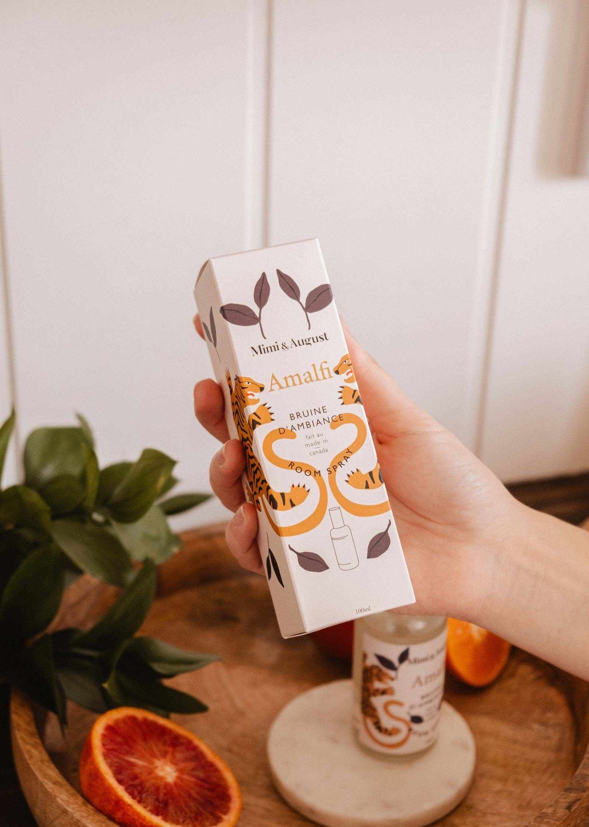 Une main tient une boîte de bruine d'ambiance Amalfi de Mimi & August au design tigré. À proximité, sur un plateau en bois avec des feuilles et une orange, la même bruine évoque une échappée méditerranéenne et une fraîcheur agrumée.