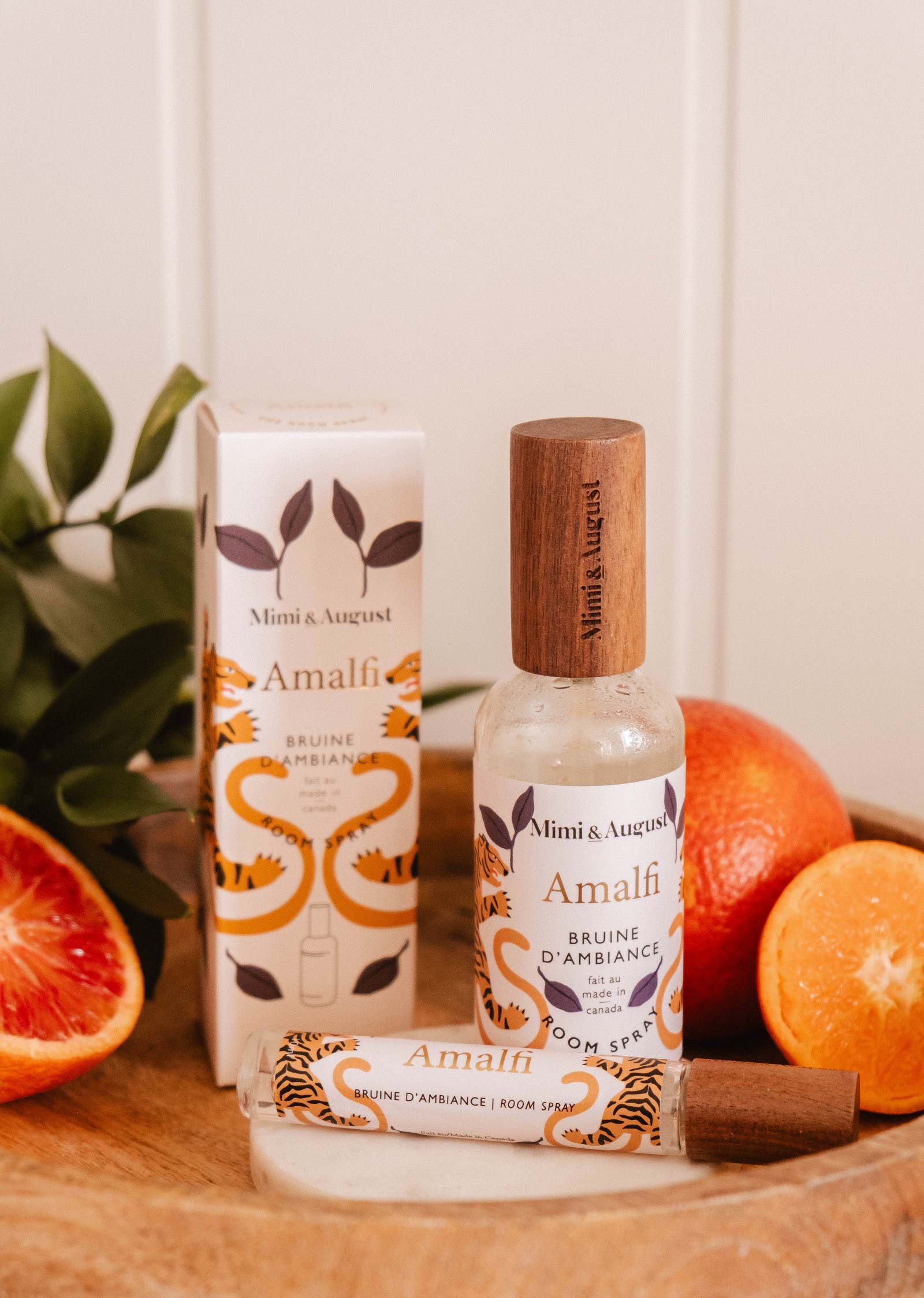 A bottle and box of Mimi & August Room spray - Amalfi are shown on a wooden surface with cut oranges and green leaves, evoking a refreshing citrus scent for a Mediterranean-inspired space.