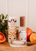 Un flacon et une boîte de Mimi & August Bruine d'ambiance - Amalfi sont présentés sur une surface en bois avec des oranges coupées et des feuilles vertes, évoquant un parfum d'agrumes rafraîchissant pour un espace d'inspiration méditerranéenne.
