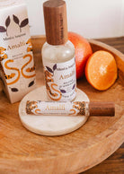 Une bouteille et un vaporisateur de bruine d'ambiance Amalfi de Mimi & August avec son emballage reposent sur un plateau en bois à côté d'une orange entière et d'une moitié d'orange, évoquant l'arôme frais d'agrumes de la Méditerranée.