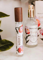 A glass Room spray Tulipa and a roller bottle, both by Mimi & August, are shown with floral designs and packaging in the background, suggesting a fresh wild berry scent.