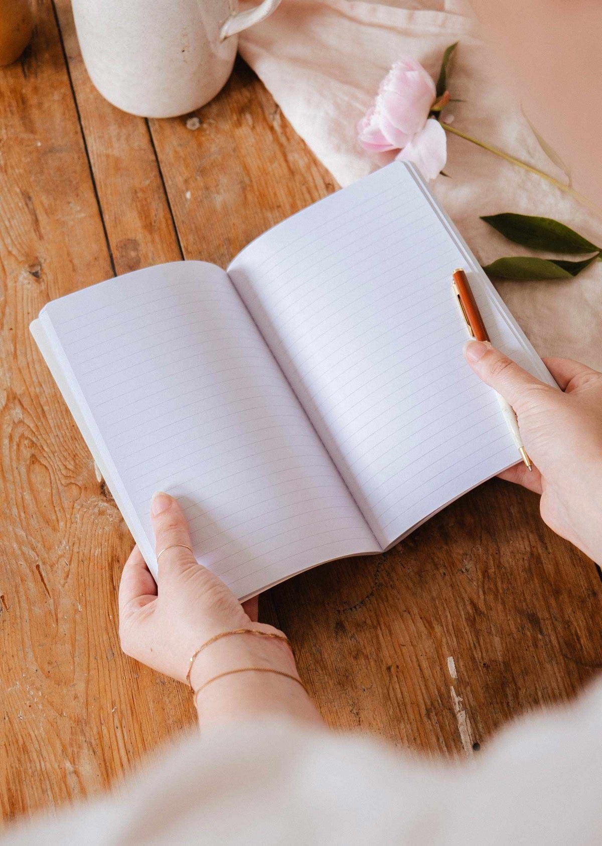 Someone holds an open Rosie - Notebook by Mimi & August with lined, floral pages and a pen on a wooden table; a pink flower and fabric are visible in the background.