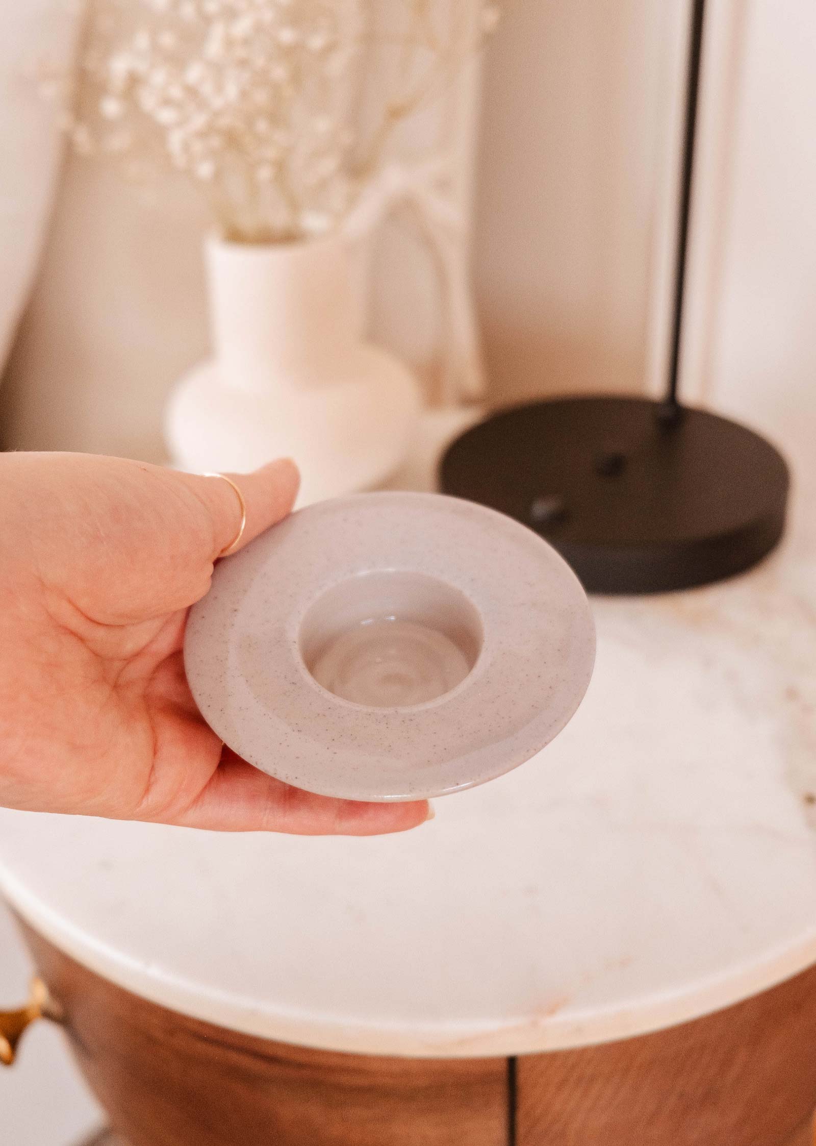 A person's hand holding a small porcelain dish with a circular indentation, possibly for holding a candle, next to a marble tabletop with decorative items in the background.