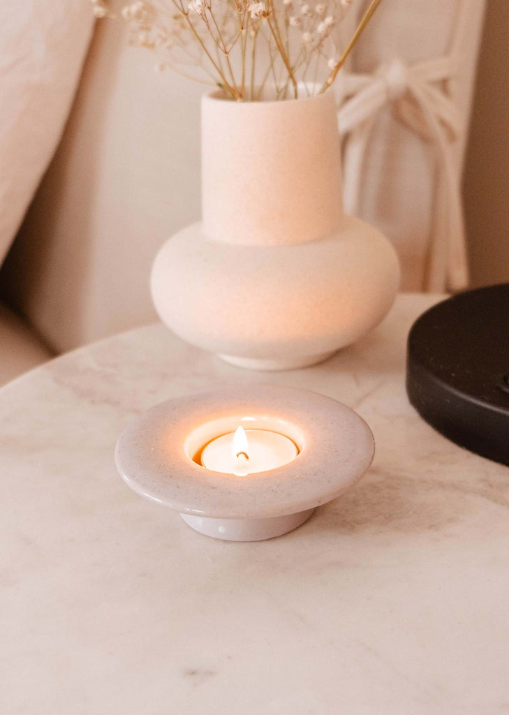 A lit candle in a Mimi & August Round Tealight Holder on a marble surface with decorative items in the background.
