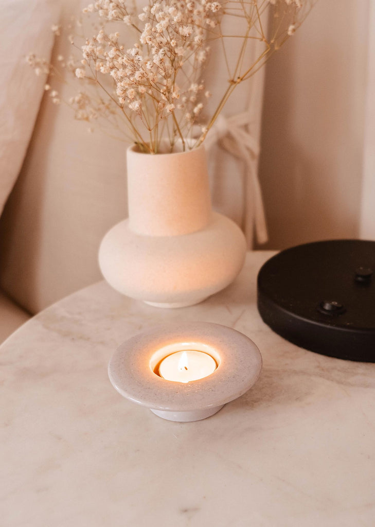A lit tealight candle in a Mimi & August Round Tealight Holder on a marble surface, accompanied by a ceramic vase with dried flowers.