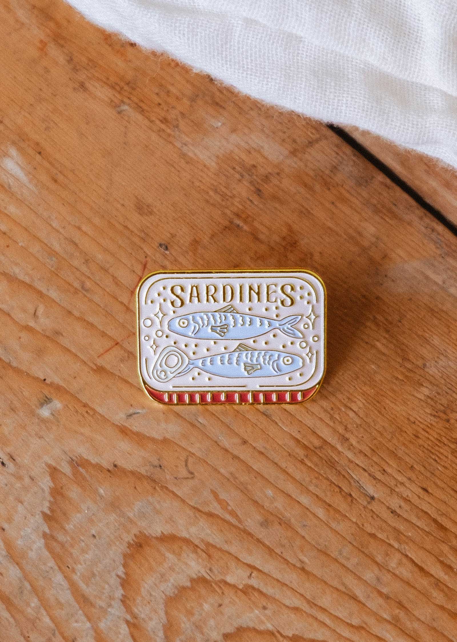 A small, rectangular Cozy Sardines Enamel Pin by Mimi & August sits on a wooden surface. Designed in Montreal, it features a sardine illustration and a red and white striped border.