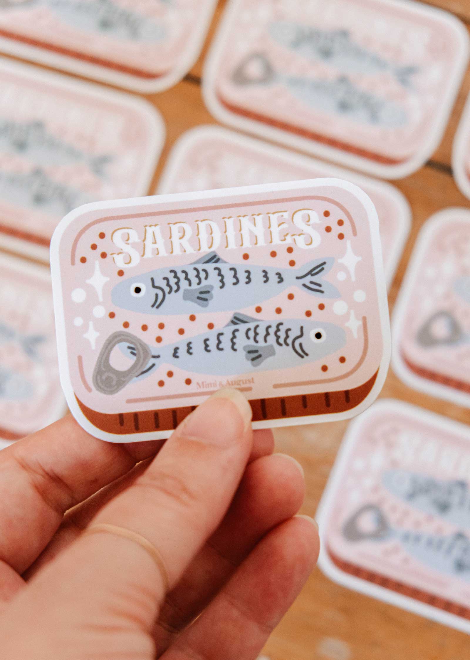 A hand holds the Cozy Sardines - Vinyl Sticker by Mimi & August, featuring an illustrated sardine can with two fish and "SARDINES" text; similar stickers are blurred in the background.