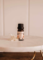 A small Scented Oil - Bloom by Mimi & August rests on a marble surface, releasing a refreshing fragrance with a small white flower beside it.