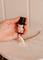 Une main tient une petite bouteille étiquetée « Huile parfumée – Bloom » de Mimi & August, libérant une fragrance rafraîchissante à côté d'une fleur blanche sur une surface en marbre.