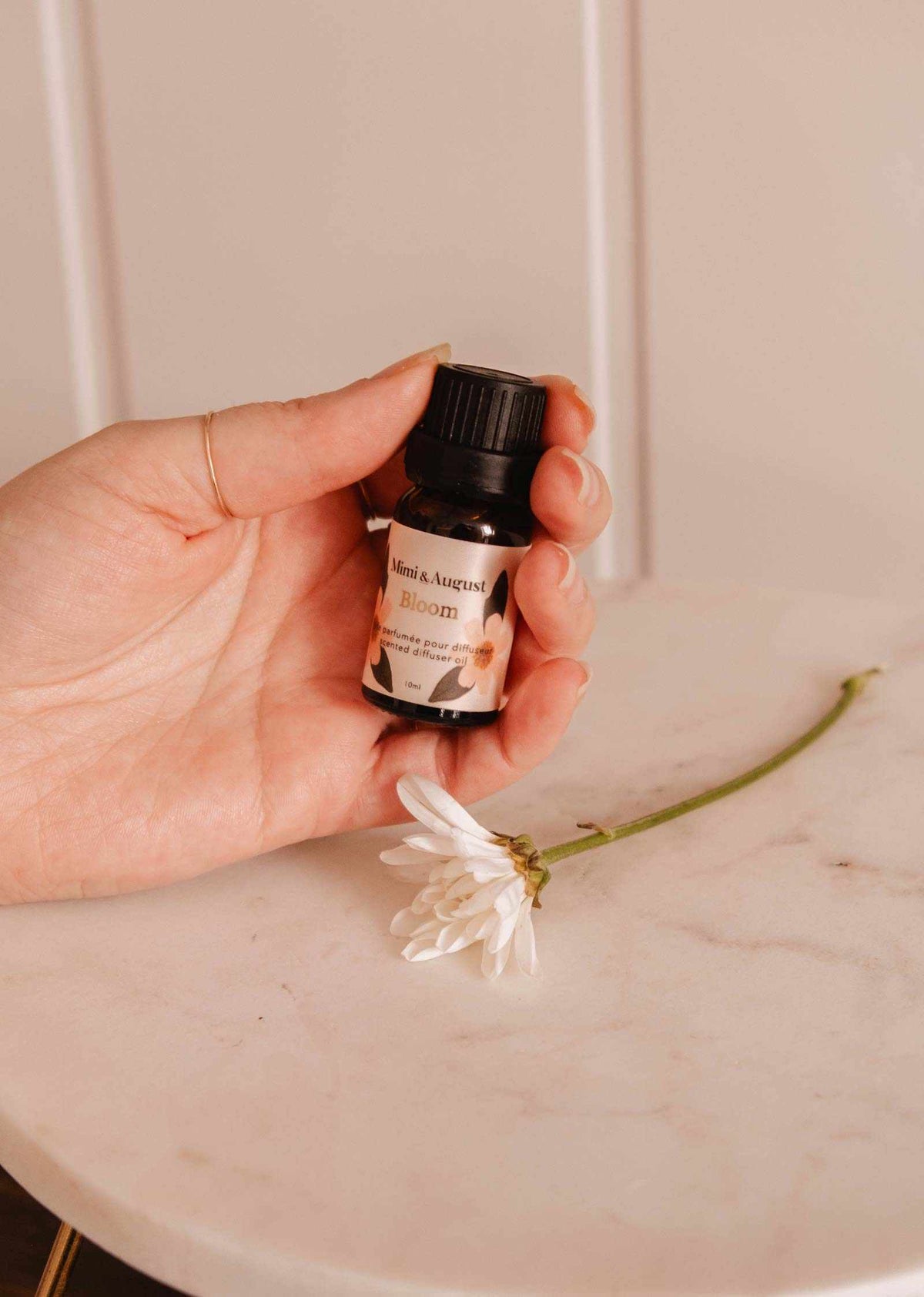 Une main tient une petite bouteille étiquetée « Huile parfumée – Bloom » de Mimi & August, libérant une fragrance rafraîchissante à côté d'une fleur blanche sur une surface en marbre.