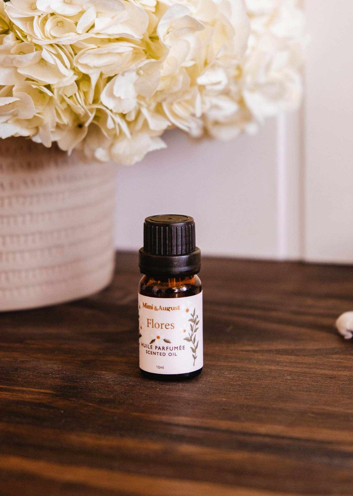 A small bottle of Mimi & August Scented Oil - Flores, featuring jasmine, lily, and mint notes, rests on a dark wooden surface before a white floral arrangement, bringing an elegant fragrance to your home.