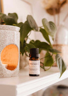 A small bottle labeled "Mimi & August Scented Oil - Lilac" is placed on a shelf beside a white ceramic oil diffuser with a lit candle inside, releasing its calming and soothing lilac scent. In the background, green leaves and neutral decor are visible.