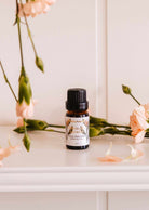 A small brown bottle of Mimi & August's Scented Oil - Oiseau du paradis, featuring a floral label, sits on a white surface surrounded by light pink flowers and green stems.