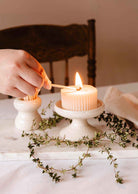 A hand lights a ribbed candle on the Mimi & August Serenity Pillar Candle Holder, which is on a marble stand with green twigs.