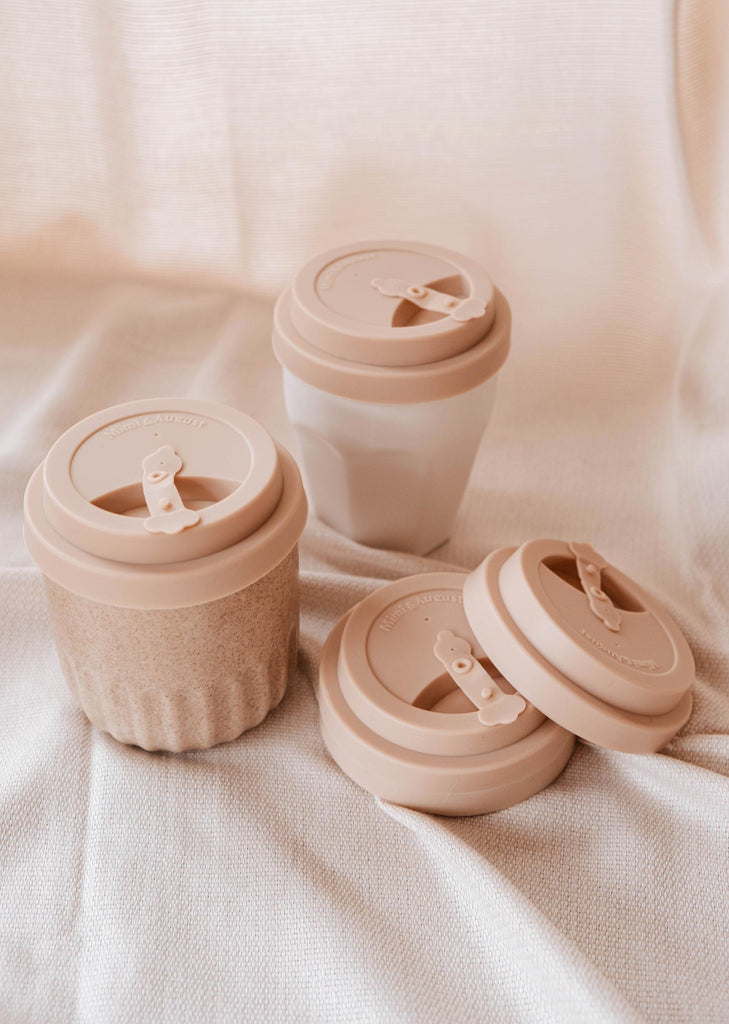 Three recyclable silicone Mimi Silicone Lid Cafe Yo Cups with lids on a bed by Mimi & August.