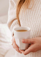 A person in a cream-colored sweater holds Mimi & August’s Soft Morning – Ceramic Cup, filled with a hot beverage.