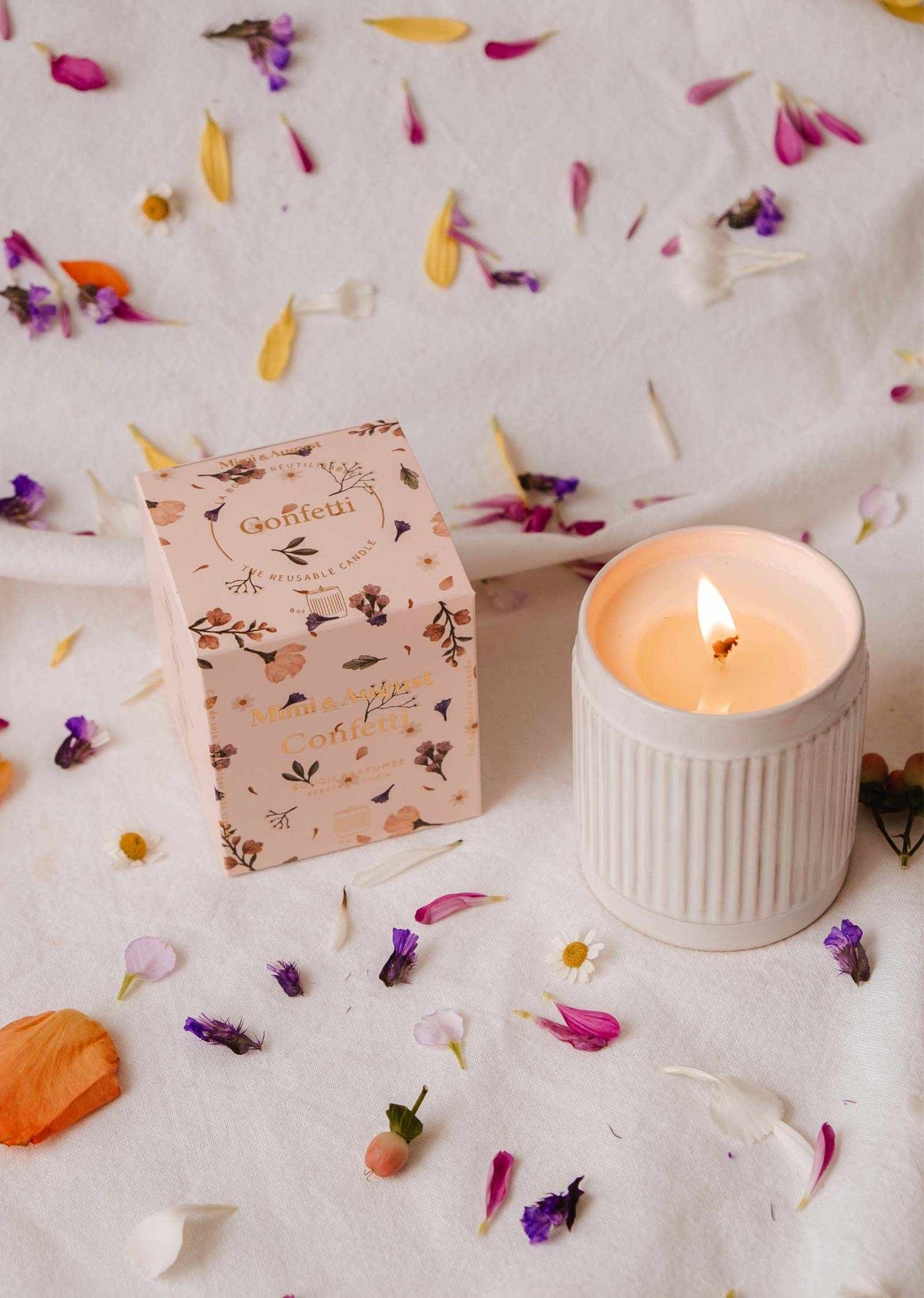 Une bougie de soya réutilisable Confetti de Mimi & August allumée dans un bougeoir cannelé repose à côté de sa boîte florale sur un tissu blanc, entourée de pétales colorés. Profitez de son ambiance chaleureuse et de son subtil parfum fruité de mandarine.