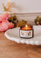 La bougie de soya Rosea 2 oz de Mimi & August brille dans son pot en verre brun sur une assiette blanche, entourée de fleurs roses et de verdure — une bougie de soya aussi belle que parfumée.