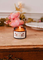 Une bougie de soya Rosea 2 oz de Mimi & August dans un pot en verre brun est allumée sur une surface en bois, avec des fleurs roses et une assiette blanche en arrière-plan.