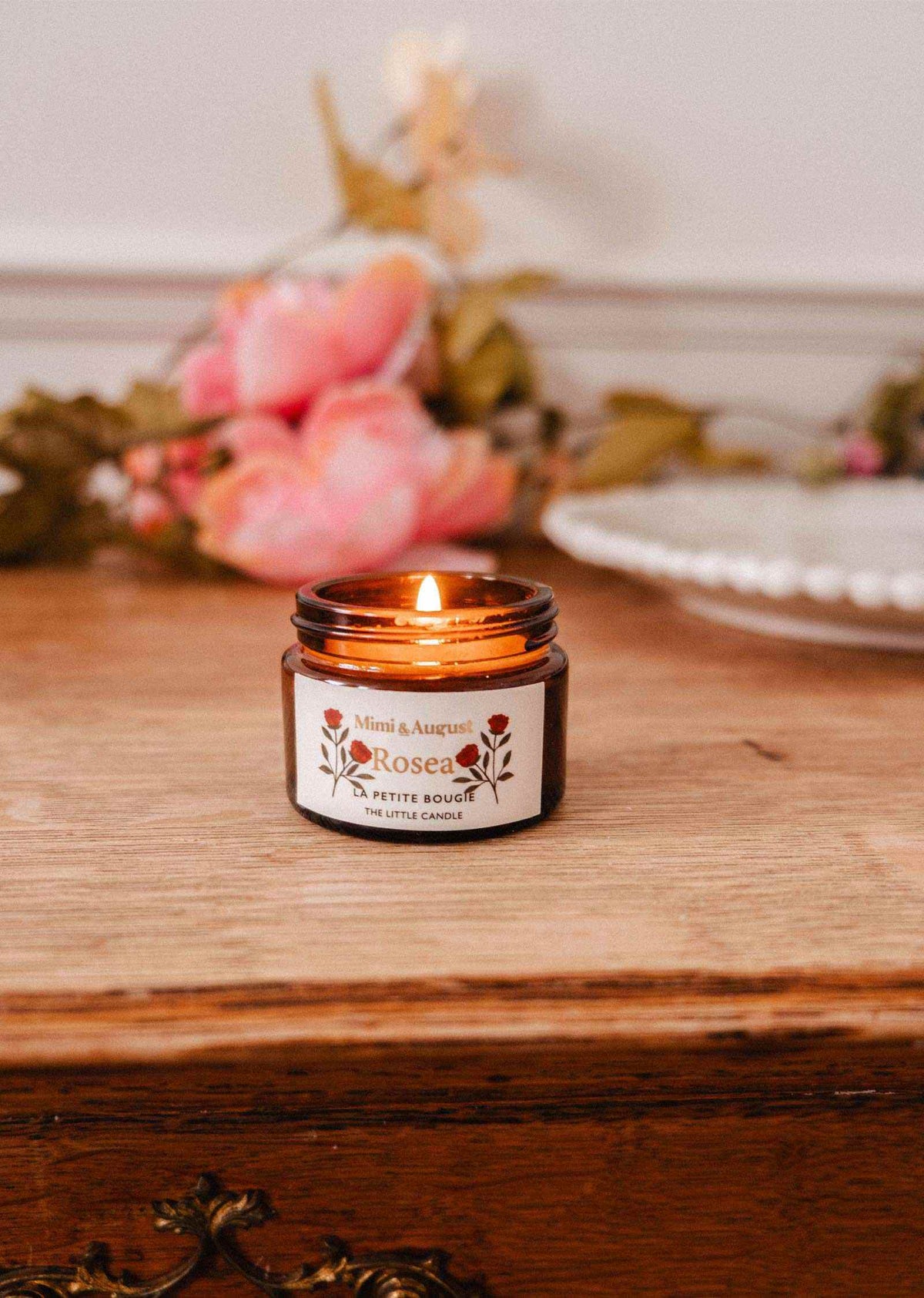 Une bougie de soya Rosea 2 oz de Mimi & August dans un pot en verre brun est allumée sur une surface en bois, avec des fleurs roses et une assiette blanche en arrière-plan.