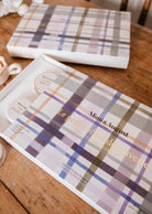 Une boîte à motif tartan portant l'inscription « Mimi & August — Crée ton kit découverte », ouverte sur une table en bois, présentant un kit découverte avec des lampions ronds en cire de soja naturelle.