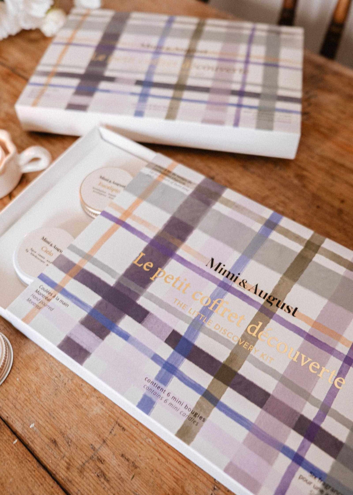 Une boîte à motif tartan portant l'inscription « Mimi & August — Crée ton kit découverte », ouverte sur une table en bois, présentant un kit découverte avec des lampions ronds en cire de soja naturelle.
