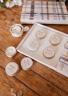 Le kit découverte personnalisable de Mimi & August comprend des contenants ronds en métal étiquetés, déposés à côté d'une boîte à motif tartan ouverte sur une table en bois, avec une bougie de soja coulée à la main et des fleurs — votre échantillonneur de bougies de soja parfait.