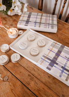 Le kit découverte personnalisable de Mimi & August, présenté dans une boîte cadeau ouverte avec des petits contenants ronds et un couvercle à motif tartan, posé sur une table en bois aux côtés d'une bougie allumée et de fleurs.