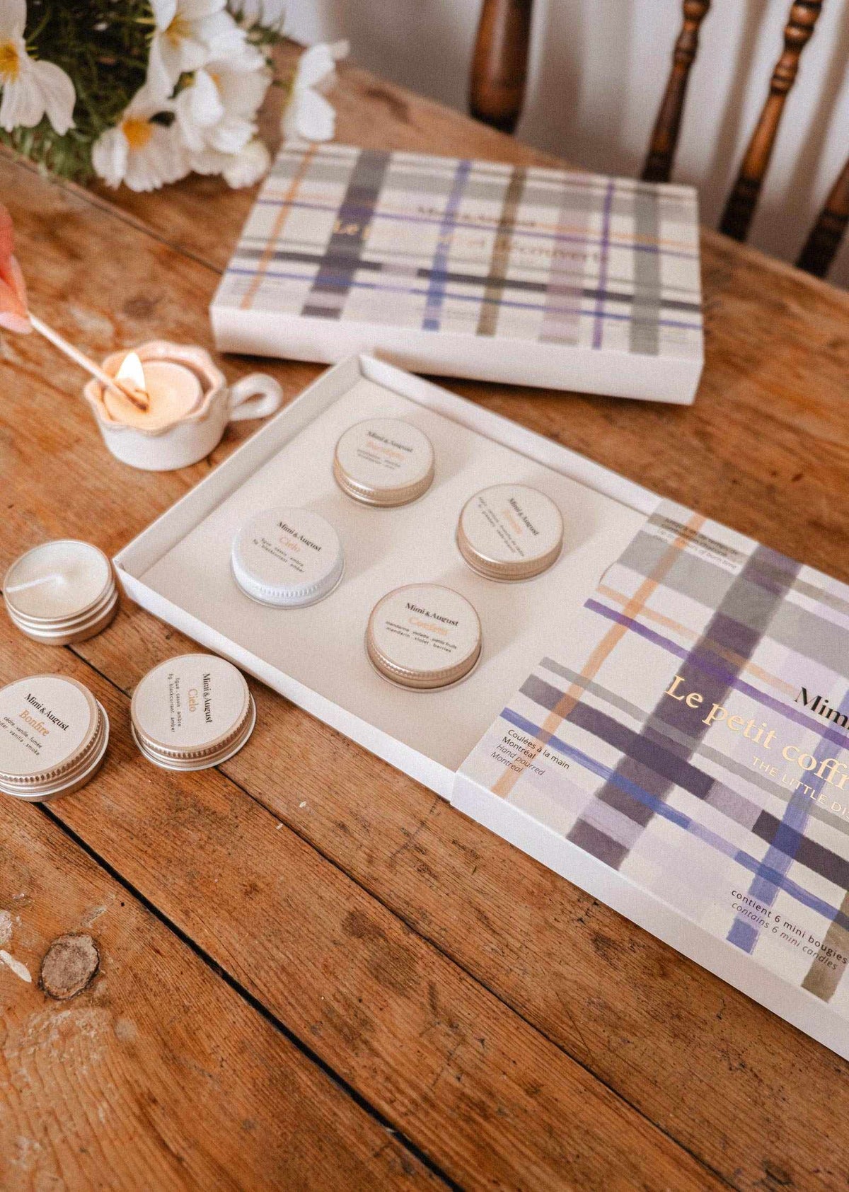 Le kit découverte personnalisable de Mimi & August, présenté dans une boîte cadeau ouverte avec des petits contenants ronds et un couvercle à motif tartan, posé sur une table en bois aux côtés d'une bougie allumée et de fleurs.