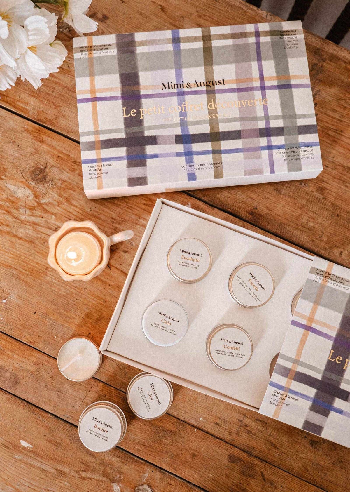 Une boîte cadeau à motif tartan portant l'inscription « Crée ton kit découverte » de Mimi & August, ouverte sur une table en bois, dévoilant six petits lampions de cire de soja. Une bougie allumée et des marguerites ajoutent du charme à la composition.