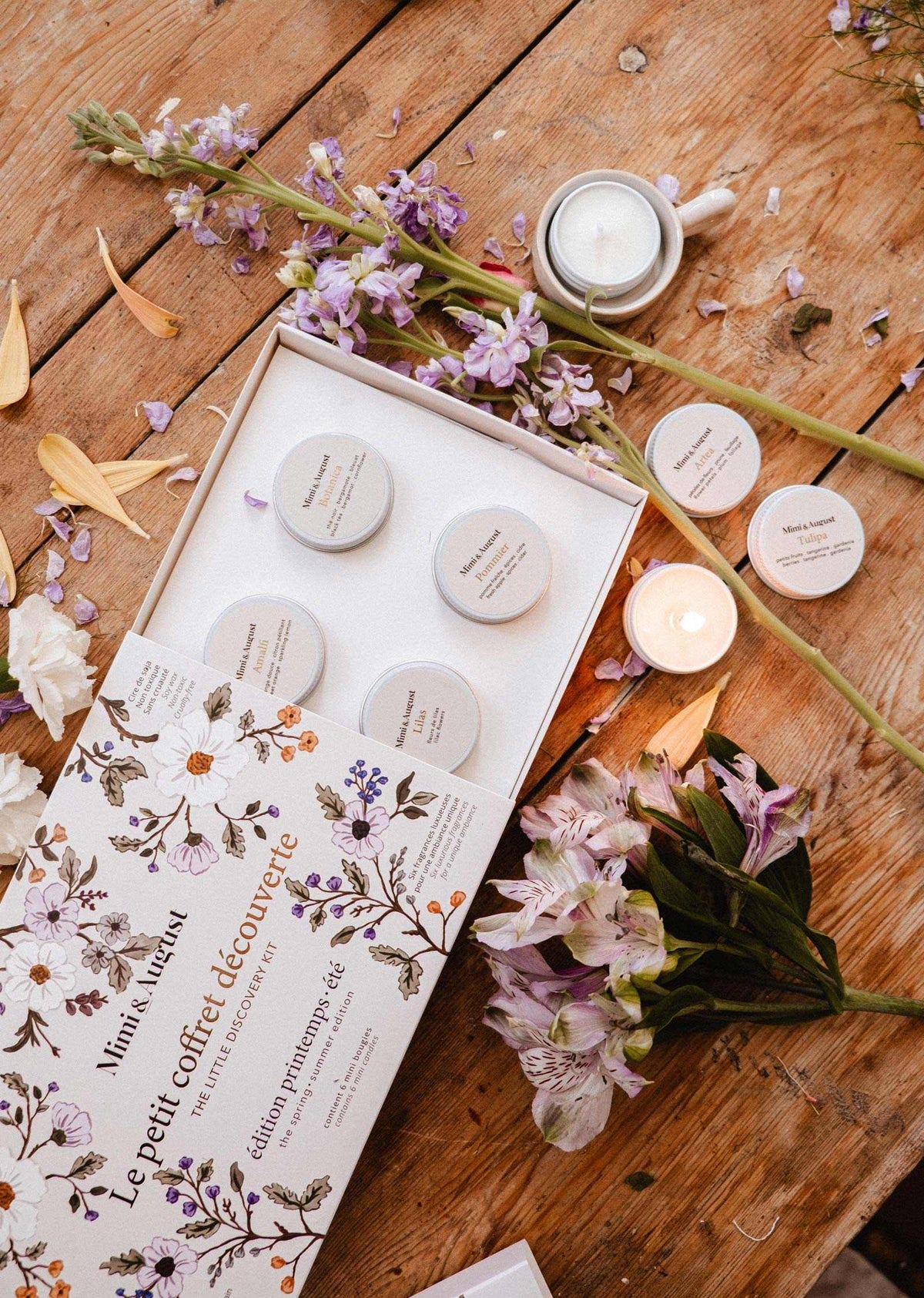 Le Kit Découverte Printemps/Été de Mimi & August, présentant cinq petits contenants, est exposé ouvert sur une table en bois avec un mini lampion en cire de soya, des fleurs violettes et blanches, des bougies et des pétales de fleurs.