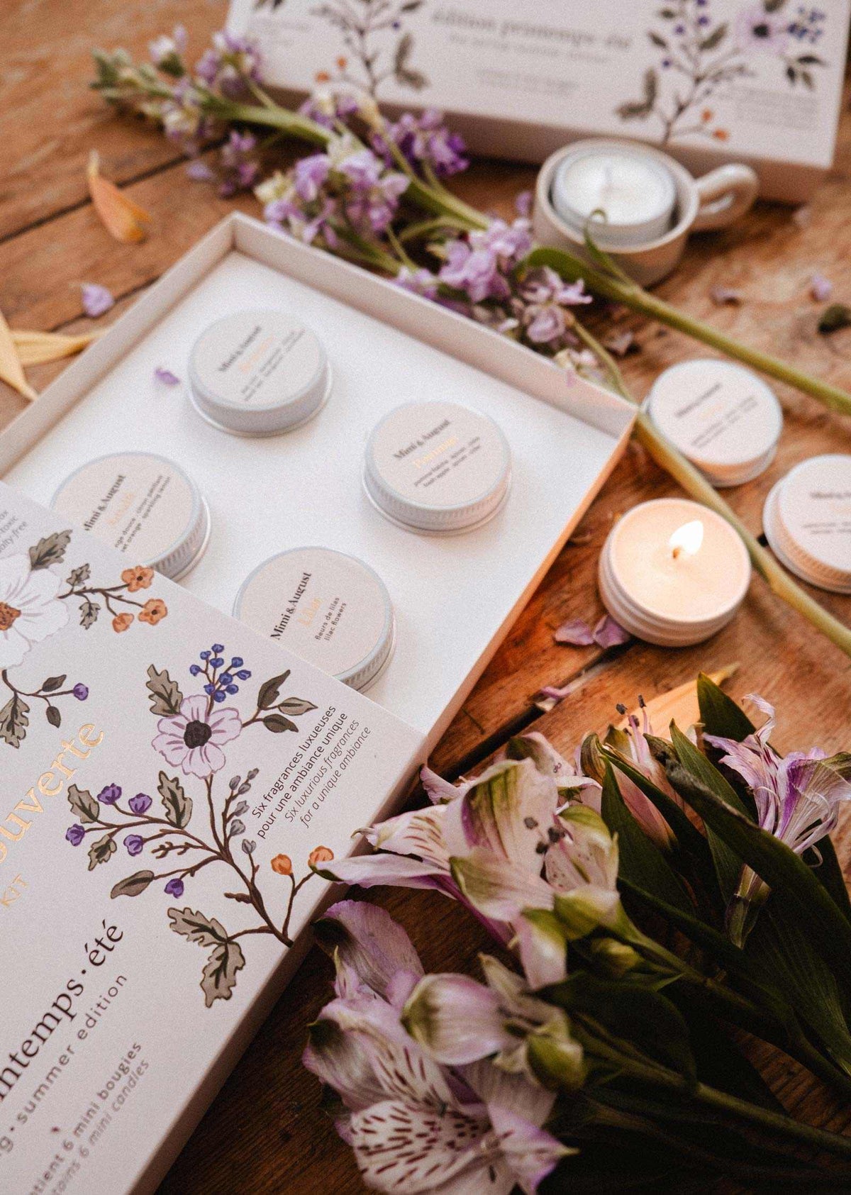 Le Kit Découverte Printemps/Été de Mimi & August présente un coffret floral ouvert avec de petits pots étiquetés et un coffret de lampions en cire de soya, exposé parmi des fleurs, des bougies et des pétales sur une surface en bois.