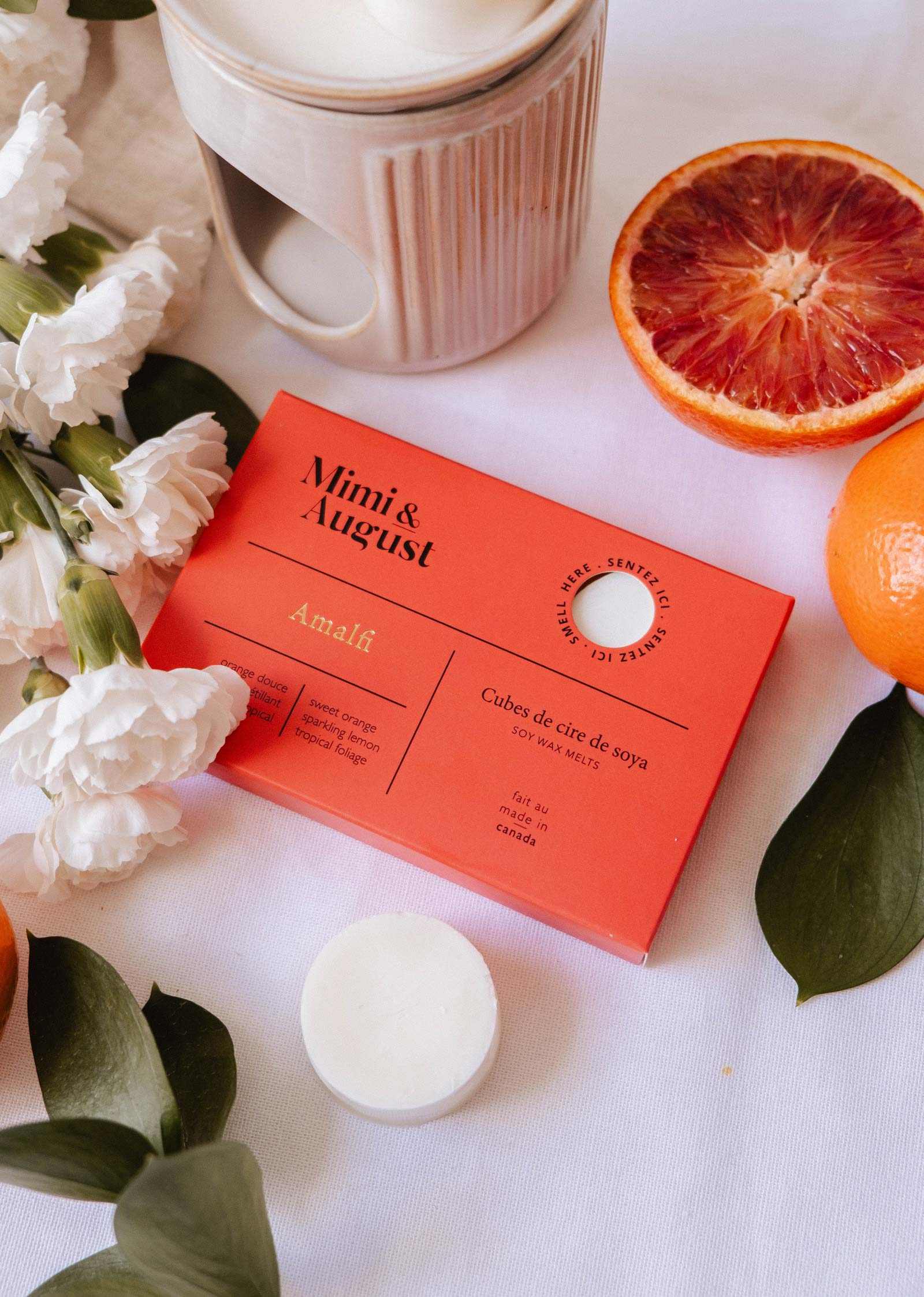 A box of Mimi & August Soy Wax Melts - Amalfi sits on a table with white flowers, orange slices, green leaves, and a ceramic wax warmer, offering a long-lasting natural fragrance from soy wax.
