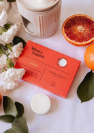 A box of Mimi & August Soy Wax Melts - Amalfi sits on a table with white flowers, orange slices, green leaves, and a ceramic wax warmer, offering a long-lasting natural fragrance from soy wax.